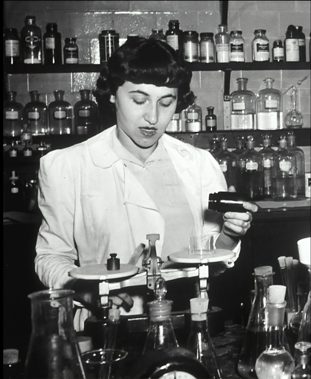 A black and white photo of a female researcher in a lab weighting a liquid on a scale.